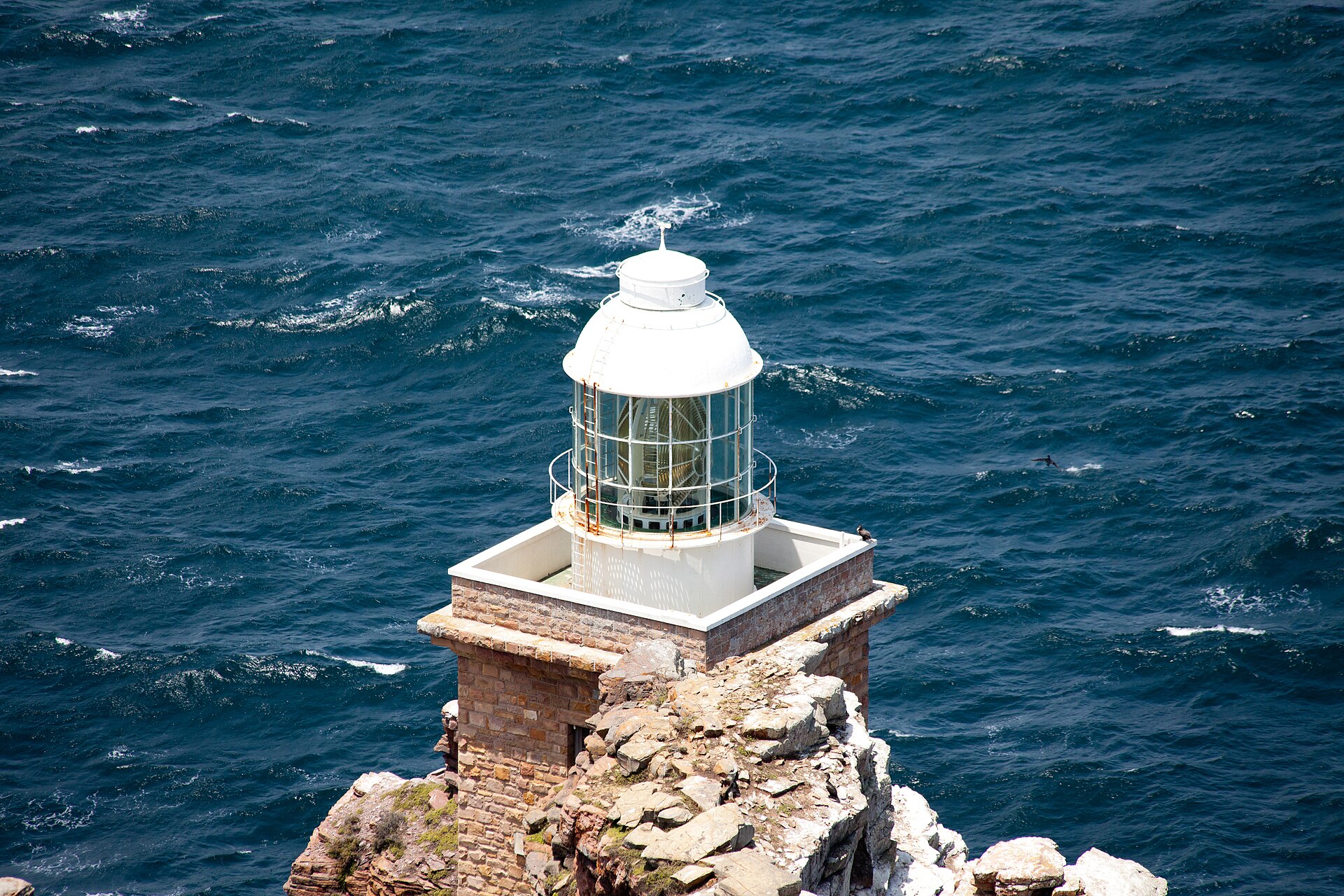 Neuer Leuchtturm von oben gesehen: weiße Laterne mit Linse auf einem gemauerten Sockel über dem dunkelblauen Meer