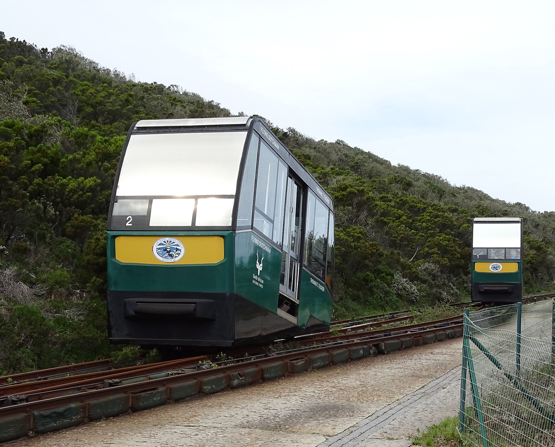 Grüner Wagen der Standseilbahn mit gelbem Schild auf der Schiene im Buschwerk, weiter hinten der zweite Wagen
