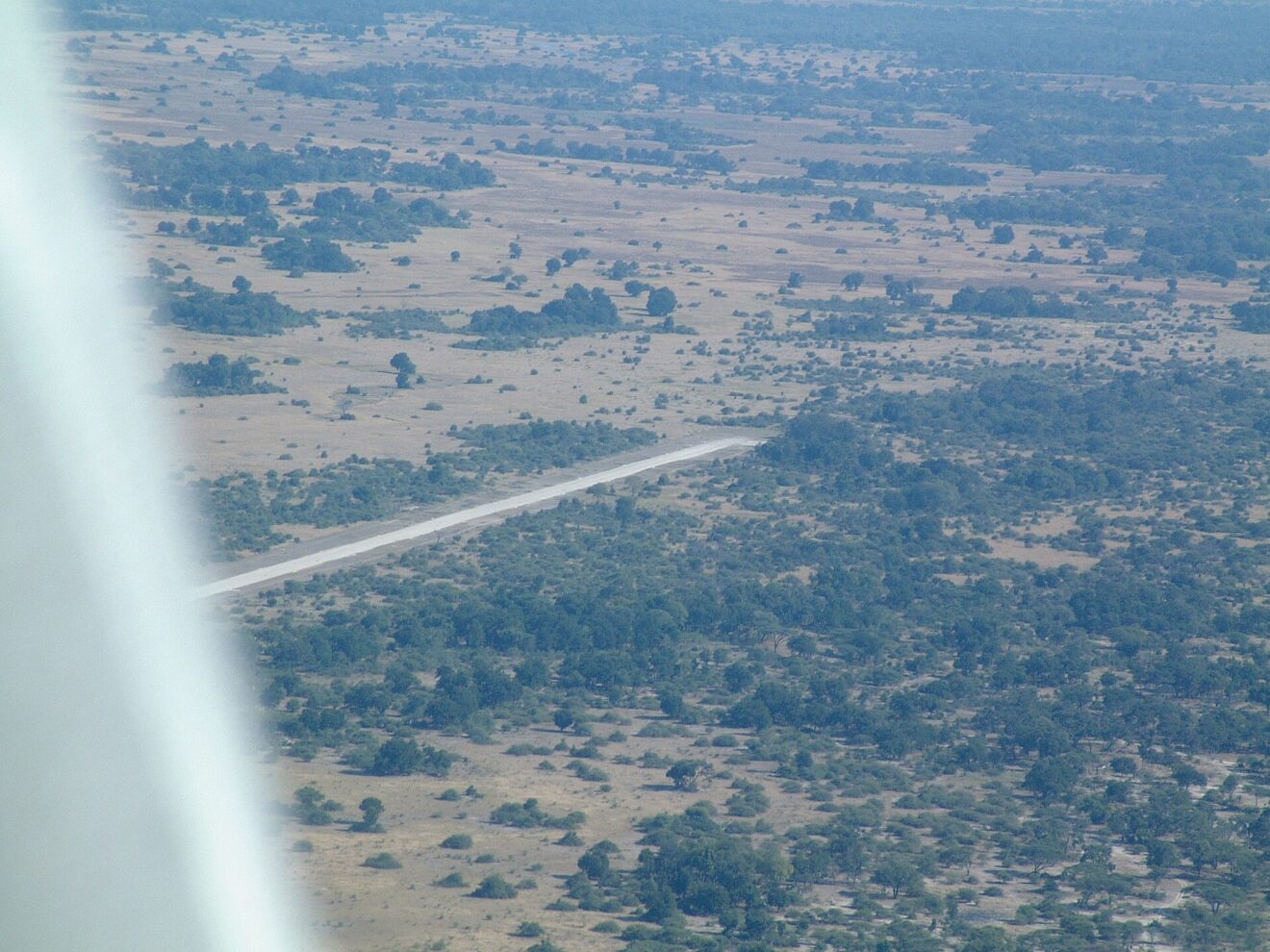 Blick aus dem Kleinflugzeug auf eine helle Landepiste zwischen Buschland und offenen Grasflächen, links die Flügelstrebe