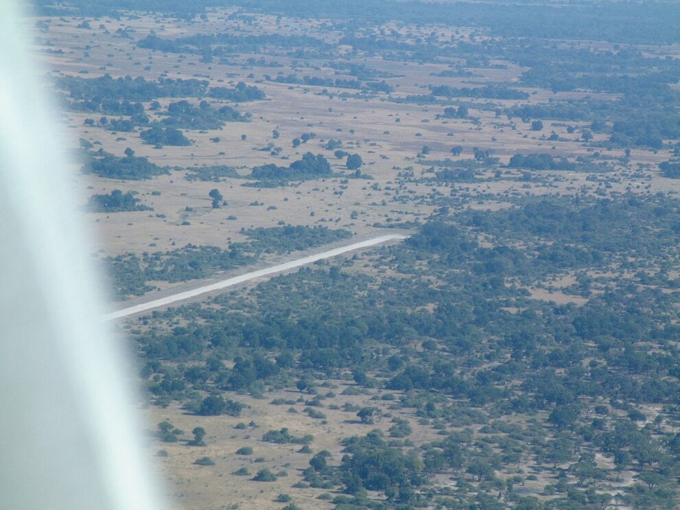 Blick aus dem Kleinflugzeug auf eine helle Landepiste zwischen Buschland und offenen Grasflächen, links die Flügelstrebe