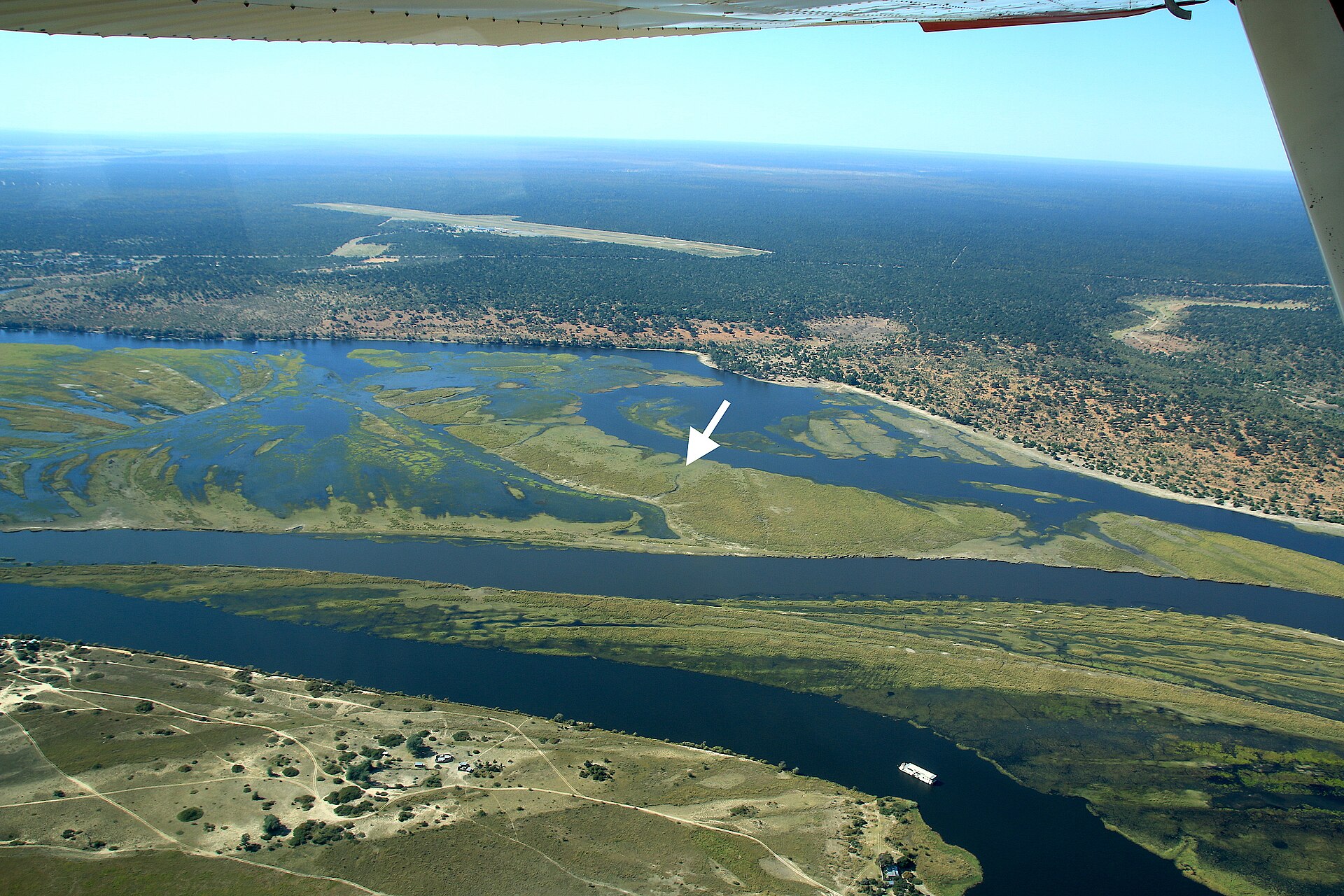 Luftbild des Chobe mit Seitenarmen und grünen Schwemmwiesen, ein weißer Pfeil zeigt auf die Insel Sedudu