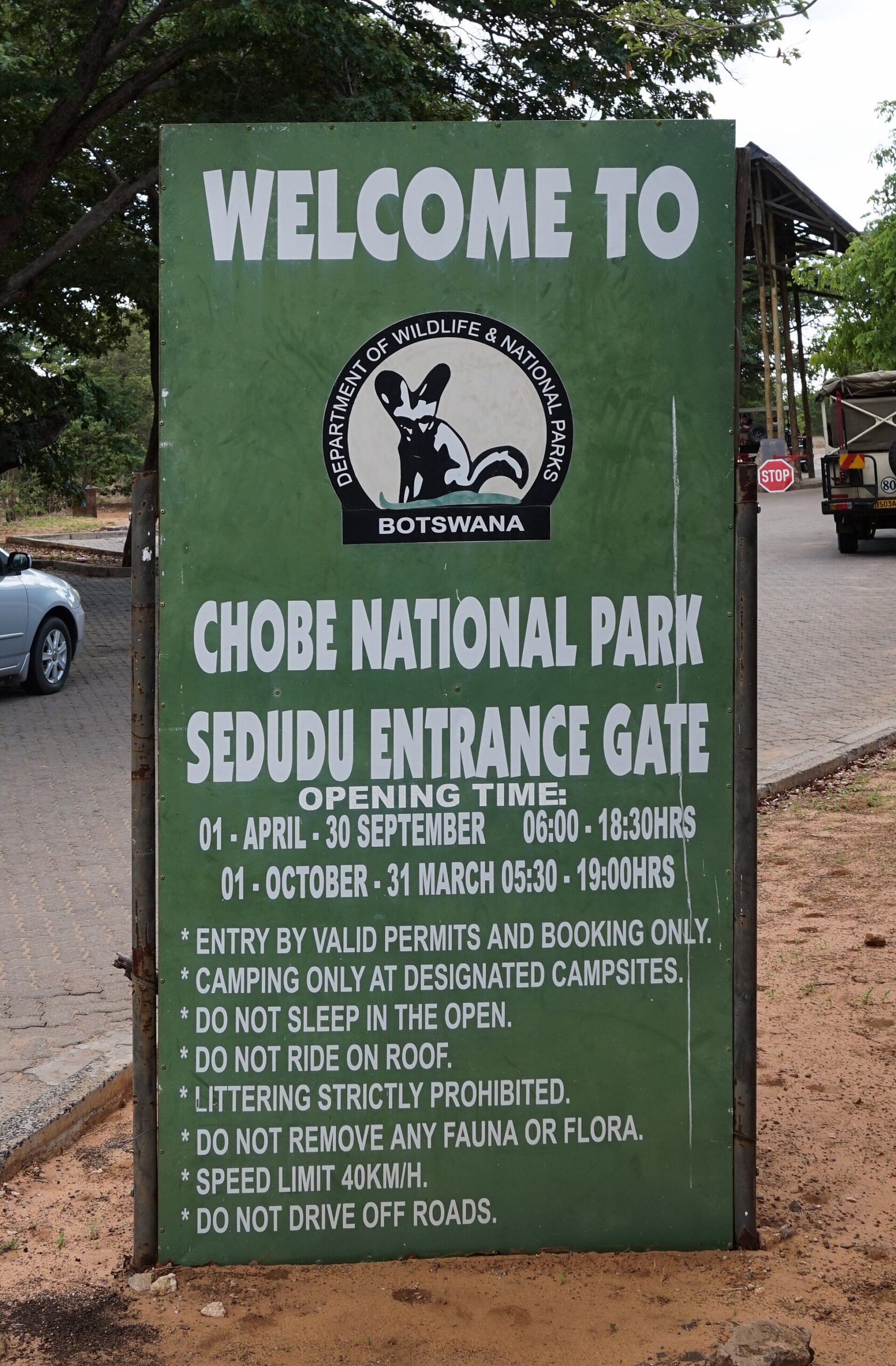 Grünes Schild „Chobe National Park, Sedudu Entrance Gate“ mit Öffnungszeiten und Parkregeln