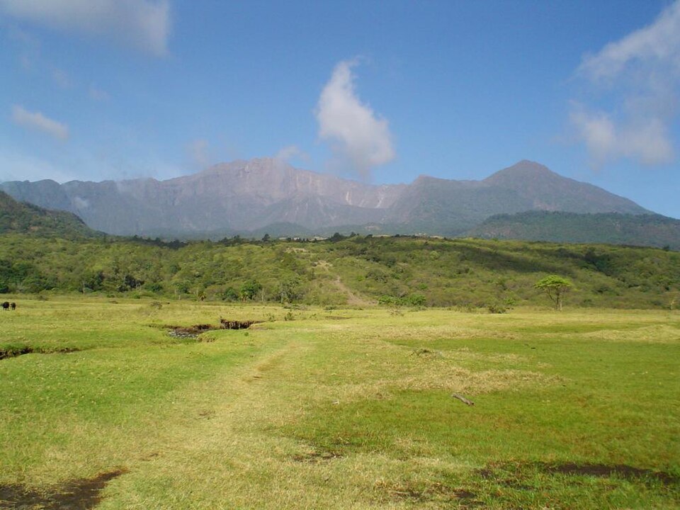 Grasland mit Pfad, dahinter bewaldete Hügel und der zerklüftete Krater des Mount Meru unter blauem Himmel