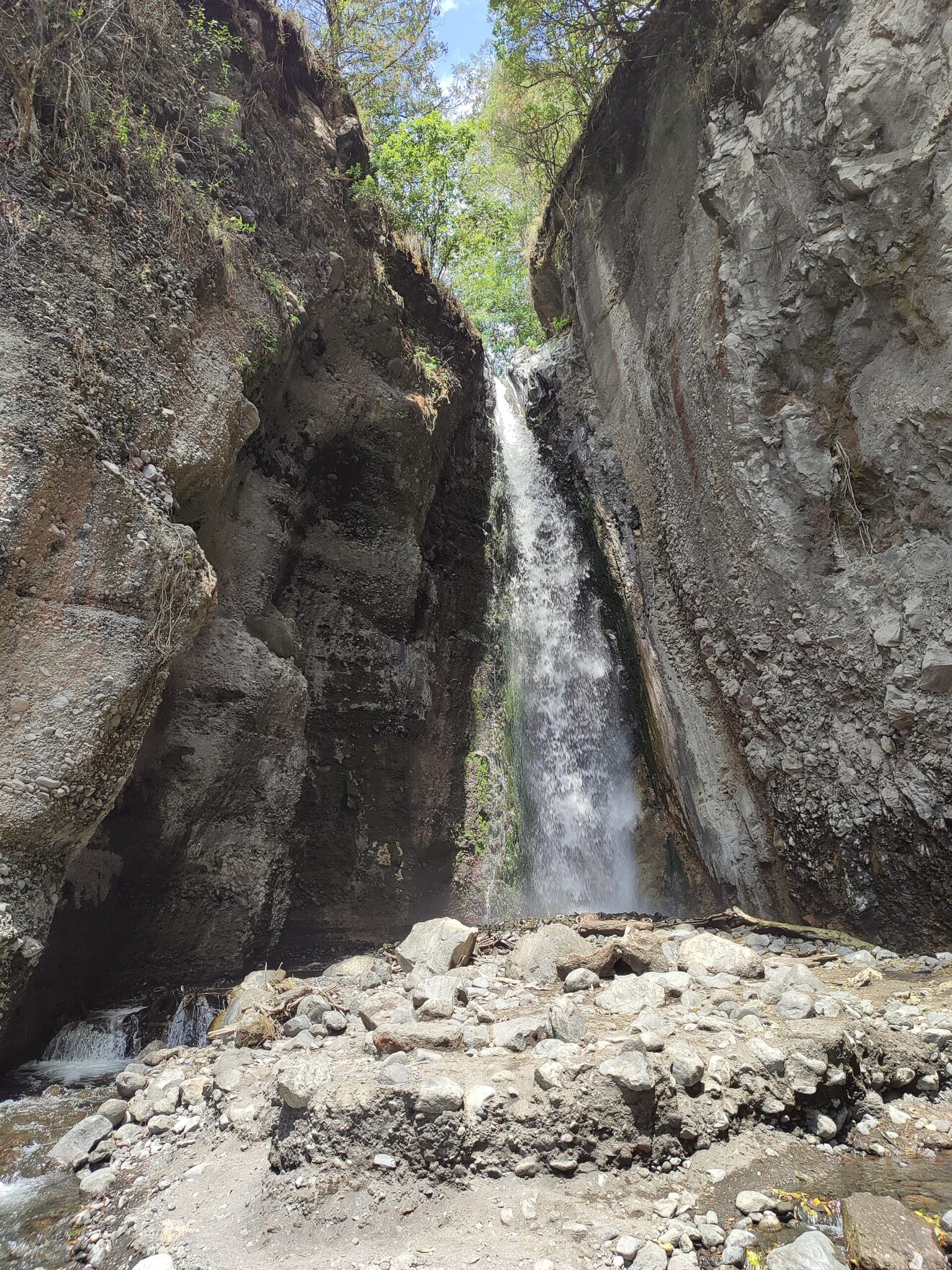 Ein schmaler Wasserfall stürzt zwischen zwei steilen grauen Felswänden in eine Schlucht mit Geröll