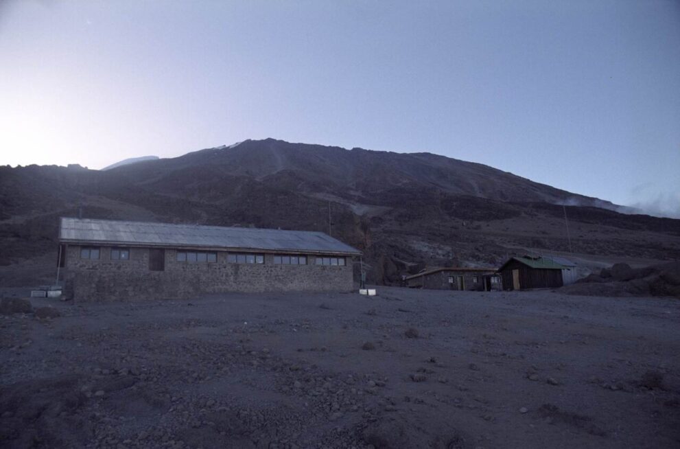 Langgestreckte Steinhütte mit Blechdach auf grauem Geröll, dahinter der dunkle Hang des Kibo in der Dämmerung
