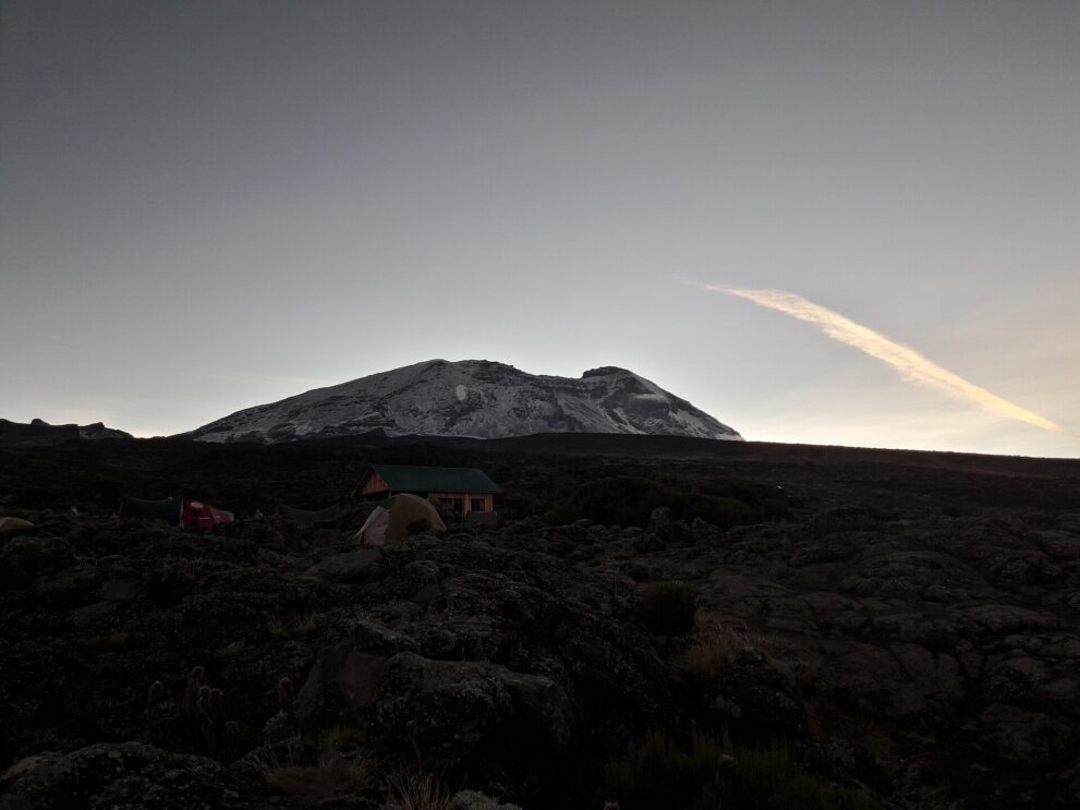 Dunkles Lavafeld mit einer Hütte und einigen Zelten, dahinter der verschneite Kibo im ersten Morgenlicht