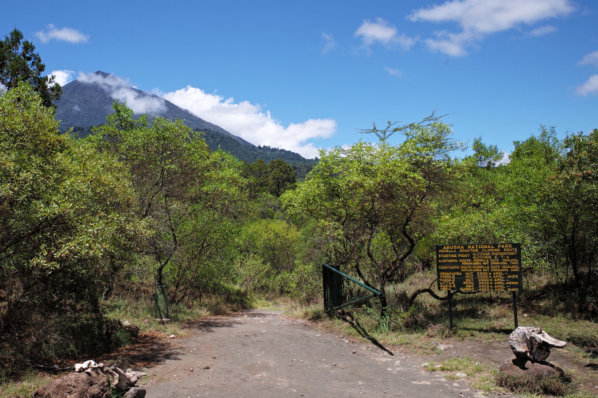 Sandweg mit offenem Schranktor, rechts eine gelbe Schautafel zur Bergroute, dahinter Buschwald und der Gipfel des Mount Meru