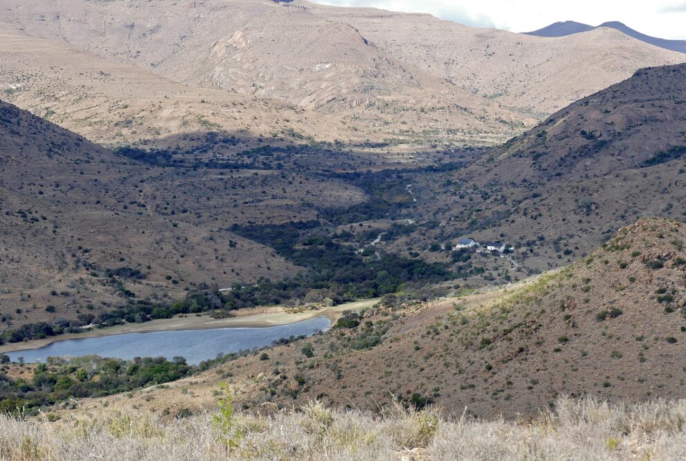 Kahle Karoo-Hügel, unten links ein Stausee, rechts davon weiße Gebäude am Hang zwischen grünem Buschwerk