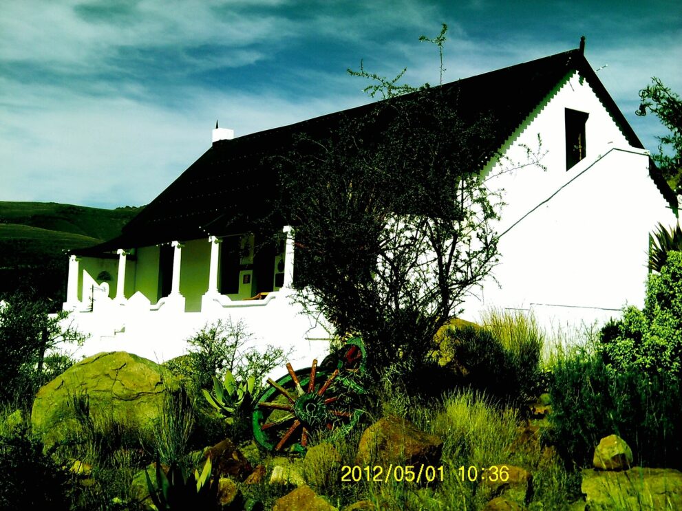 Weißes Farmhaus mit dunklem Dach, Veranda mit weißen Säulen, davor Felsen, Aloen und ein altes Wagenrad