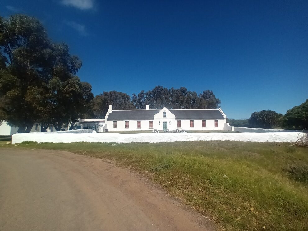 Langgestrecktes weißes Farmhaus mit Mittelgiebel hinter einer weiß gekalkten Mauer, davor eine Sandpiste, links große Bäume