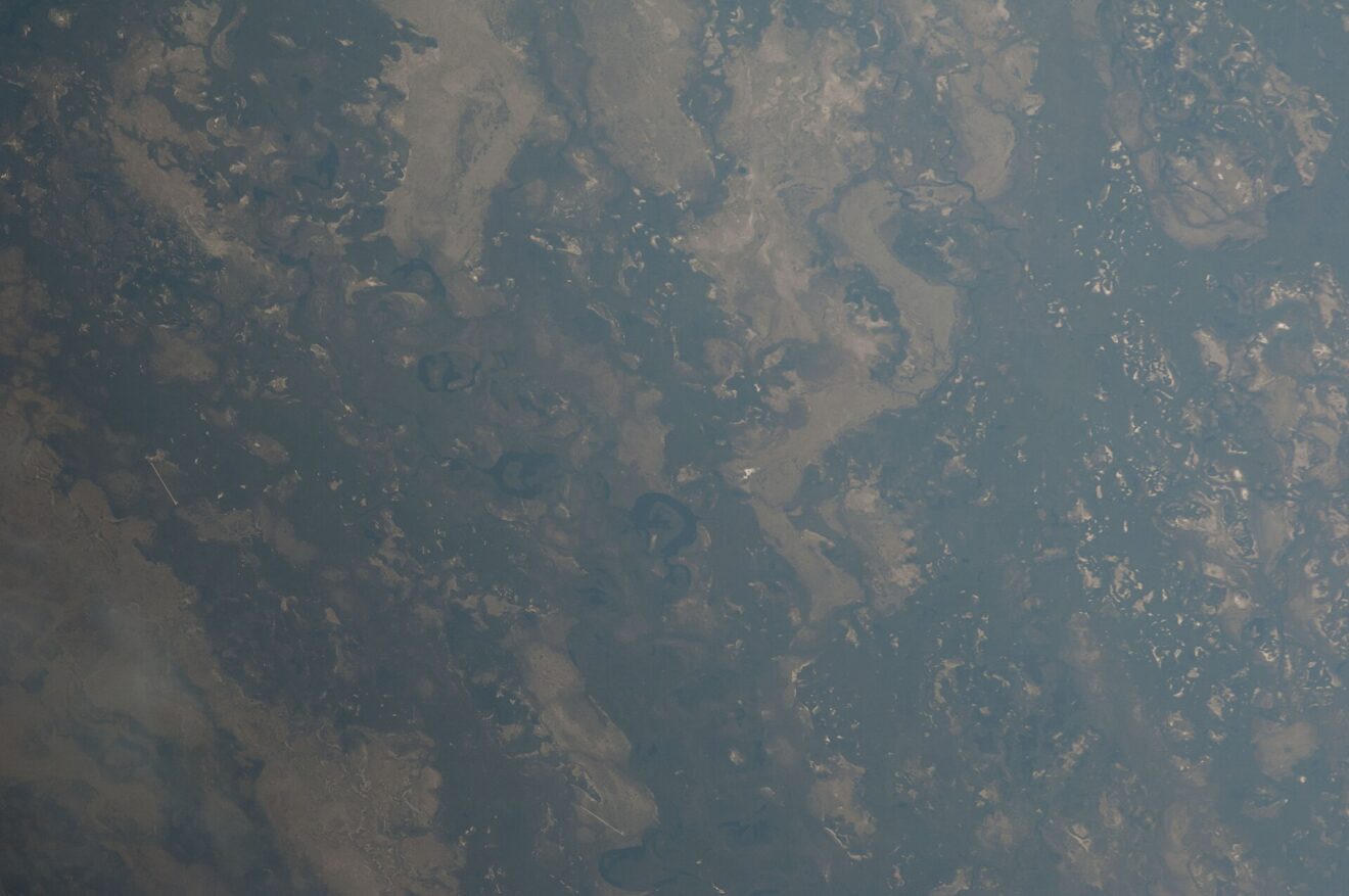 Schrägaufnahme aus dem All auf das Okavango-Delta: dunkle Wasserflächen, helle Inseln und mehrere runde, dunkle Lagunen in der Bildmitte.