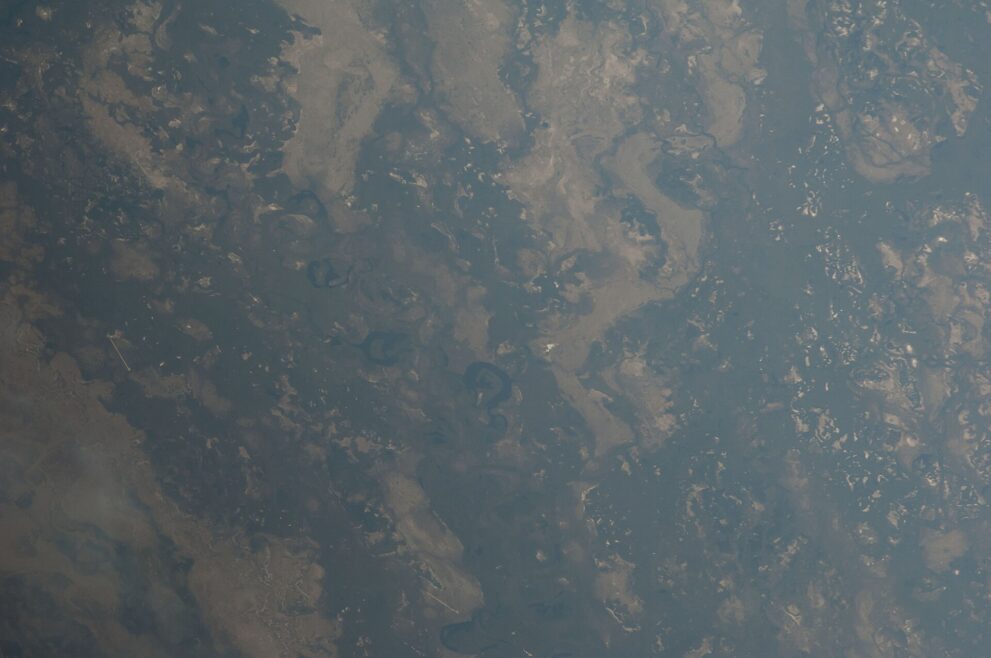 Schrägaufnahme aus dem All auf das Okavango-Delta: dunkle Wasserflächen, helle Inseln und mehrere runde, dunkle Lagunen in der Bildmitte.