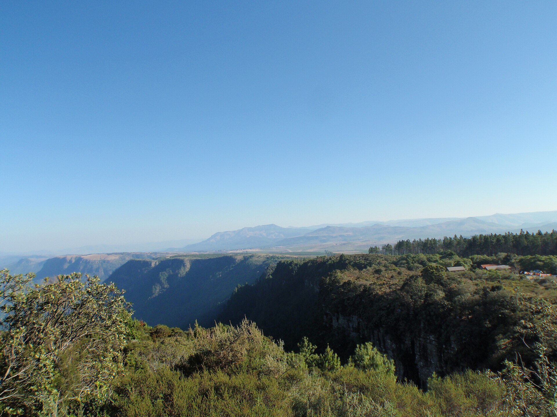 Tiefe, bewaldete Schlucht unter einer Felskante, dahinter Bergketten unter blauem Himmel, rechts am Waldrand Dächer am Parkplatz
