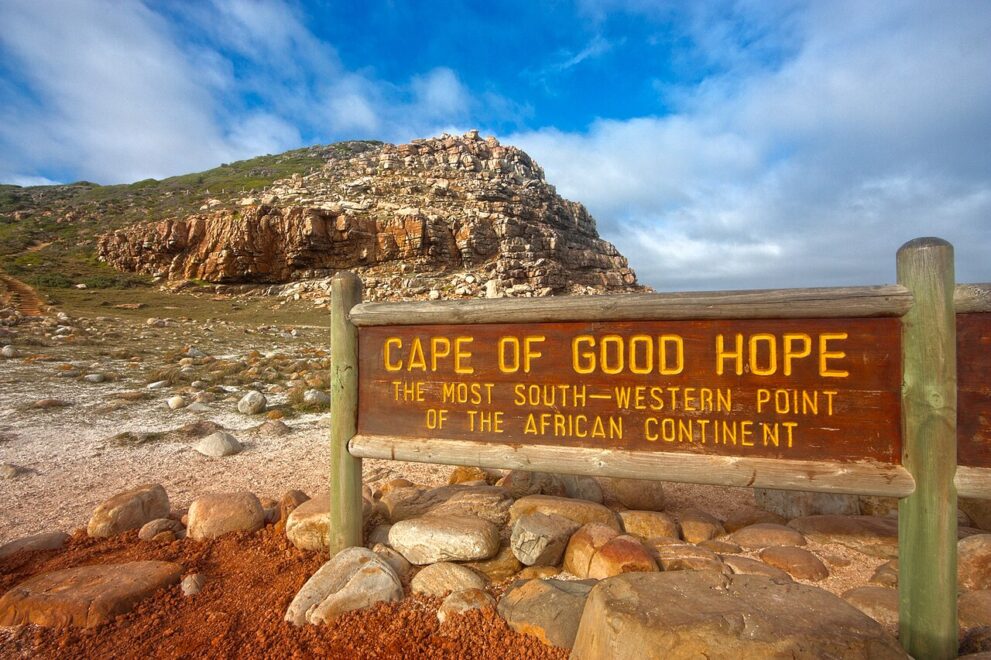 Holzschild mit der gelben Aufschrift Cape of Good Hope, the most south-western point of the African continent, dahinter ein Felshügel und Wolken