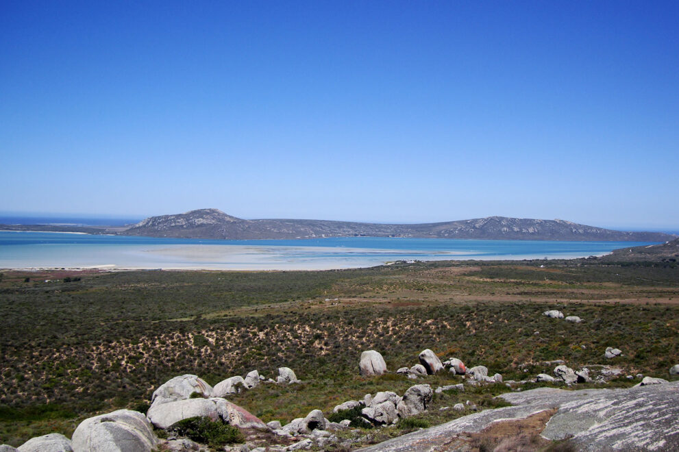 Blick über grünes Buschland auf die hellblaue Lagune mit weißen Sandbänken, dahinter die Hügel der Halbinsel und der Atlantik.