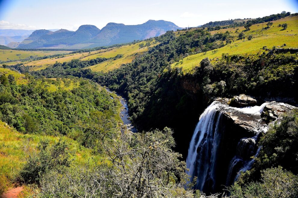 Rechts stürzt der Wasserfall über eine Felskante, links windet sich der Fluss durch eine bewaldete Schlucht, dahinter grüne Hänge und Berge.