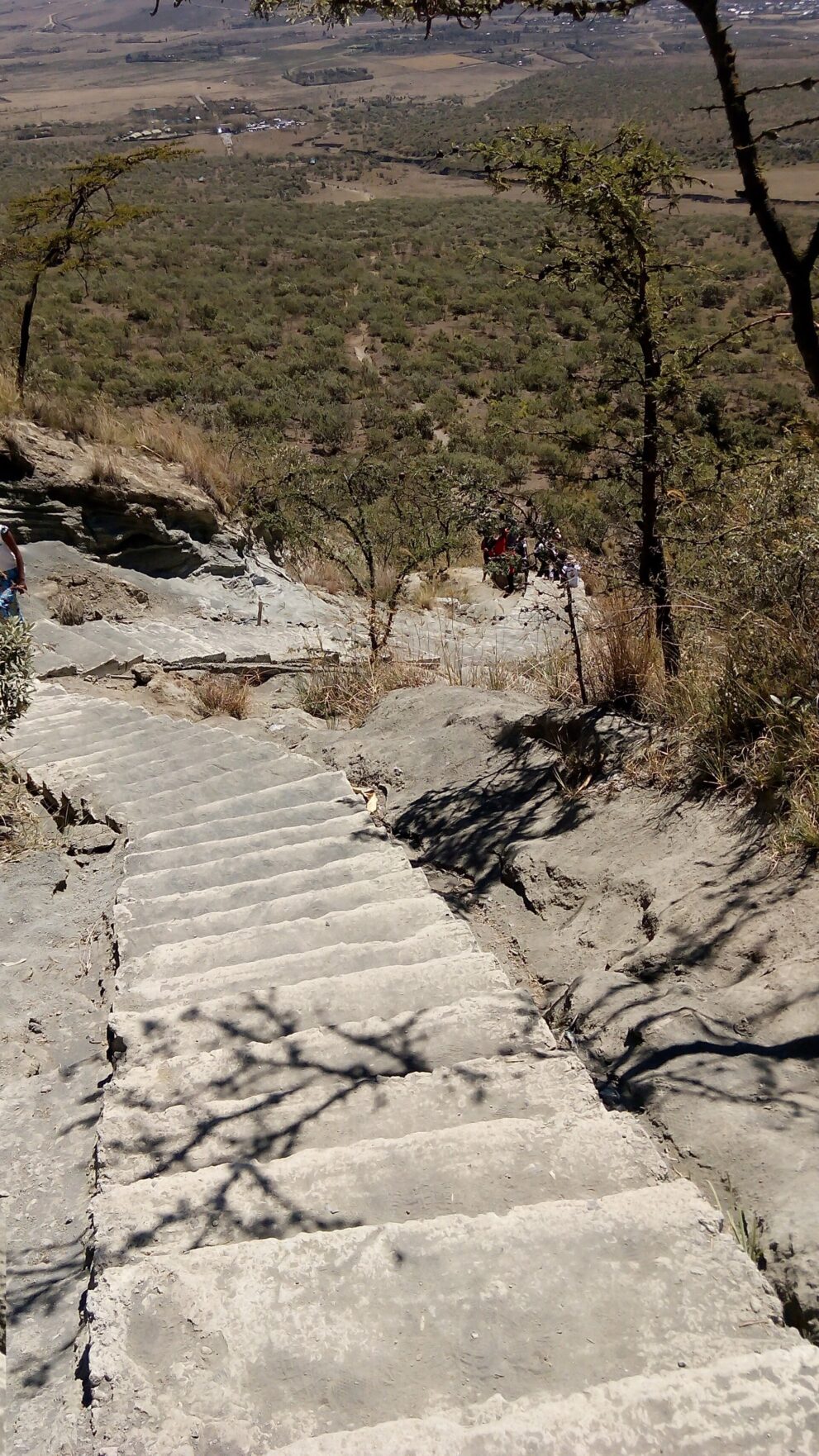 Steile Stufen aus Vulkangestein führen den Hang hinab, darunter Buschland und die Ebene des Rift Valley