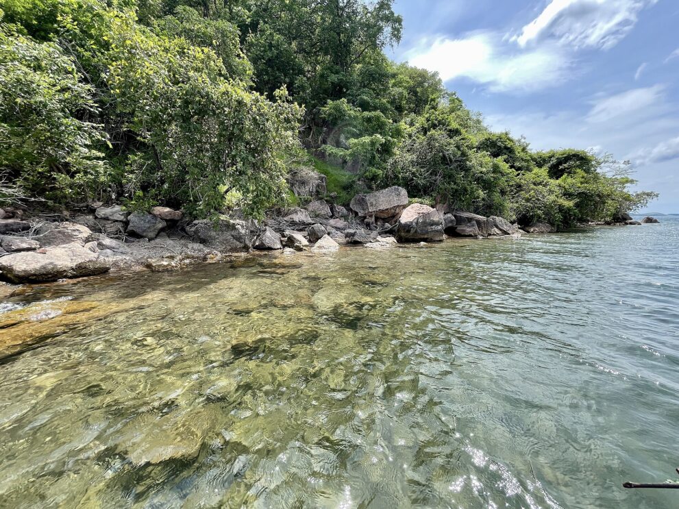 Glasklares, flaches Seewasser über hellen Steinen, am Ufer Felsblöcke und dichtes Grün