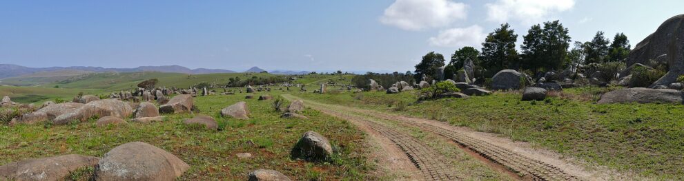 Panorama: Eine Sandpiste führt durch Grasland voller runder Granitblöcke, rechts Bäume, am Horizont blaue Berge.
