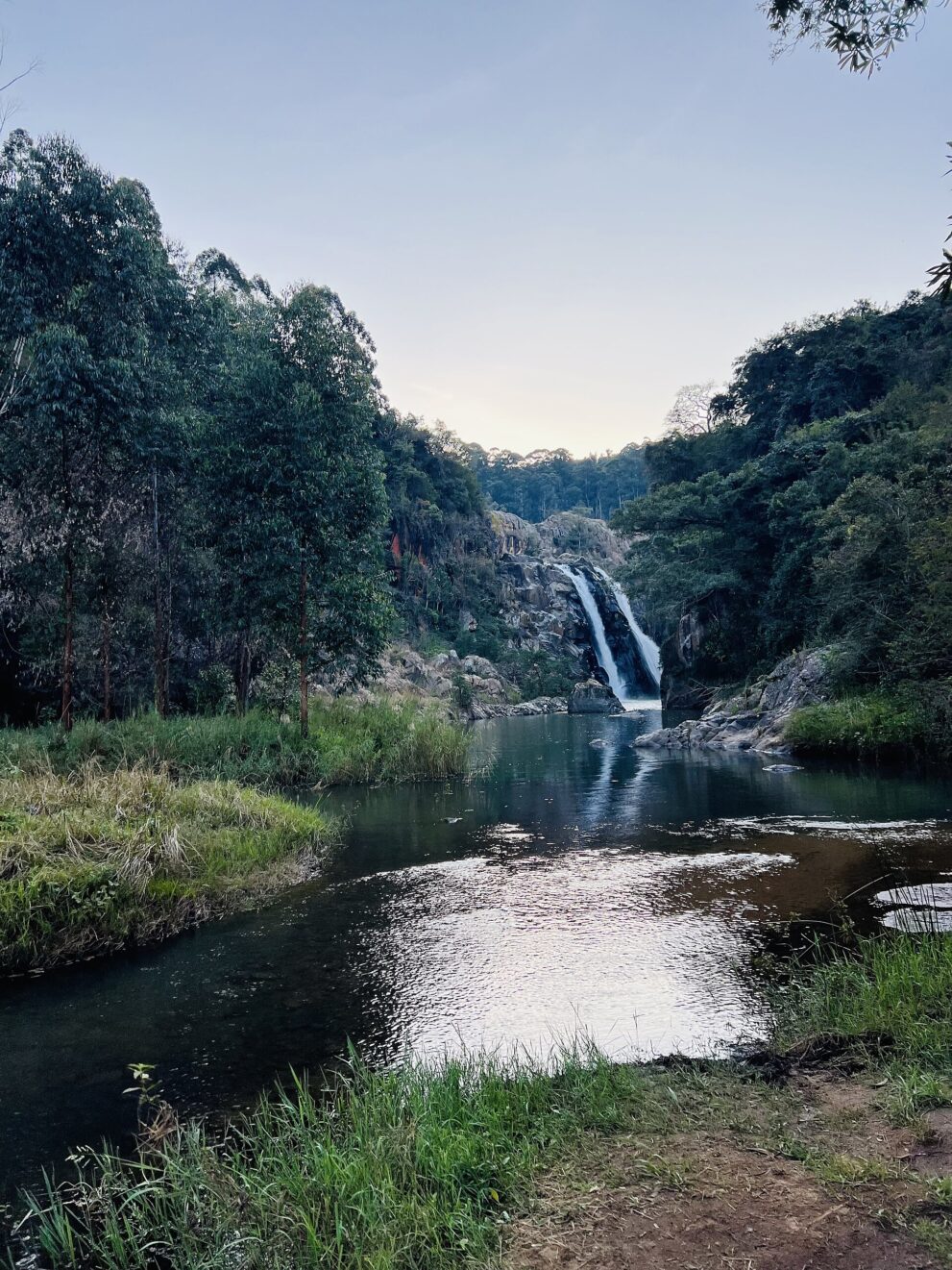 Abendlicht am Fluss: Im Hintergrund fällt der Wasserfall zwischen bewaldeten Hängen, vorn ein stilles Becken und Gras.
