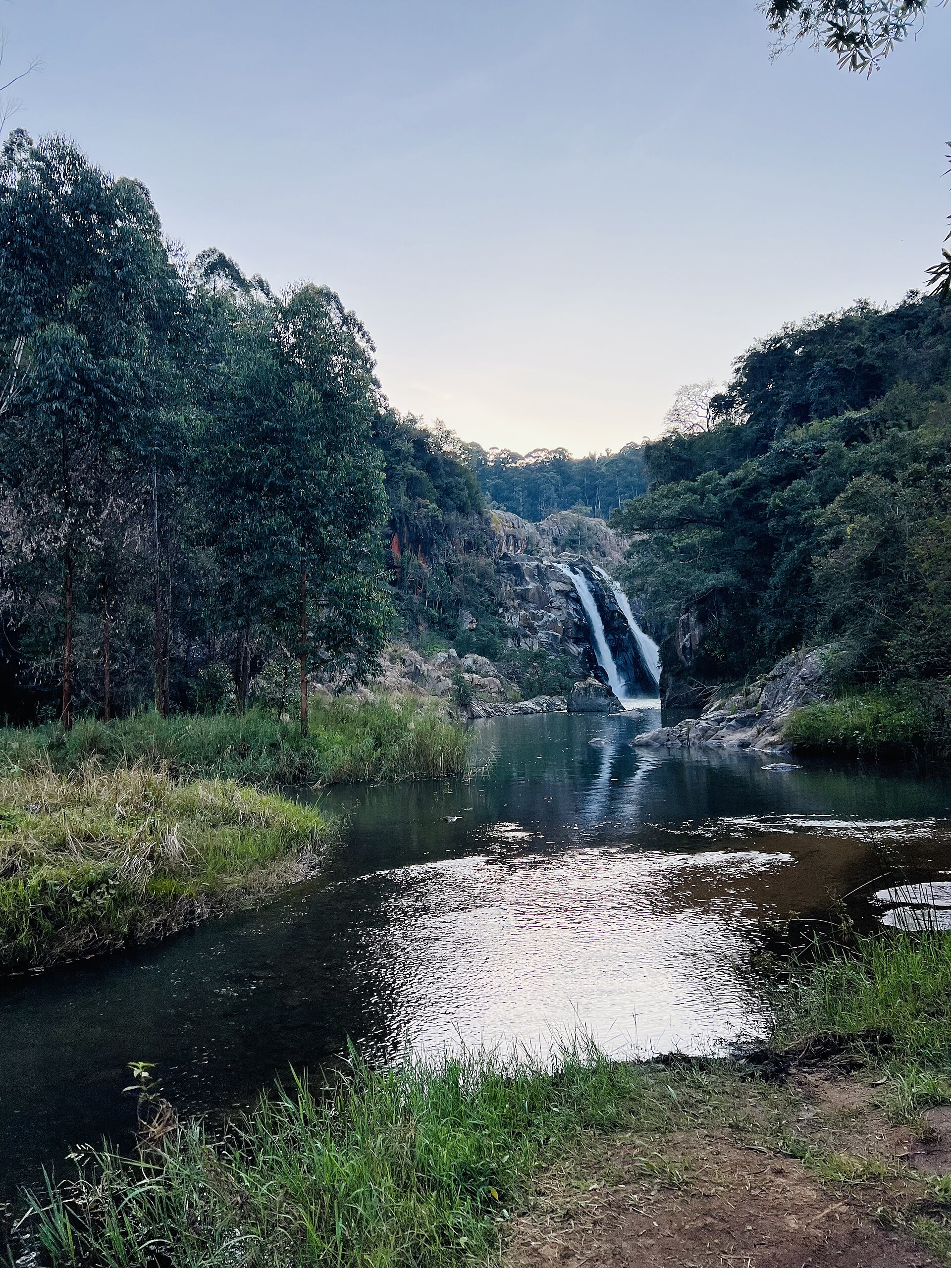 Abendlicht am Fluss: Im Hintergrund fällt der Wasserfall zwischen bewaldeten Hängen, vorn ein stilles Becken und Gras.