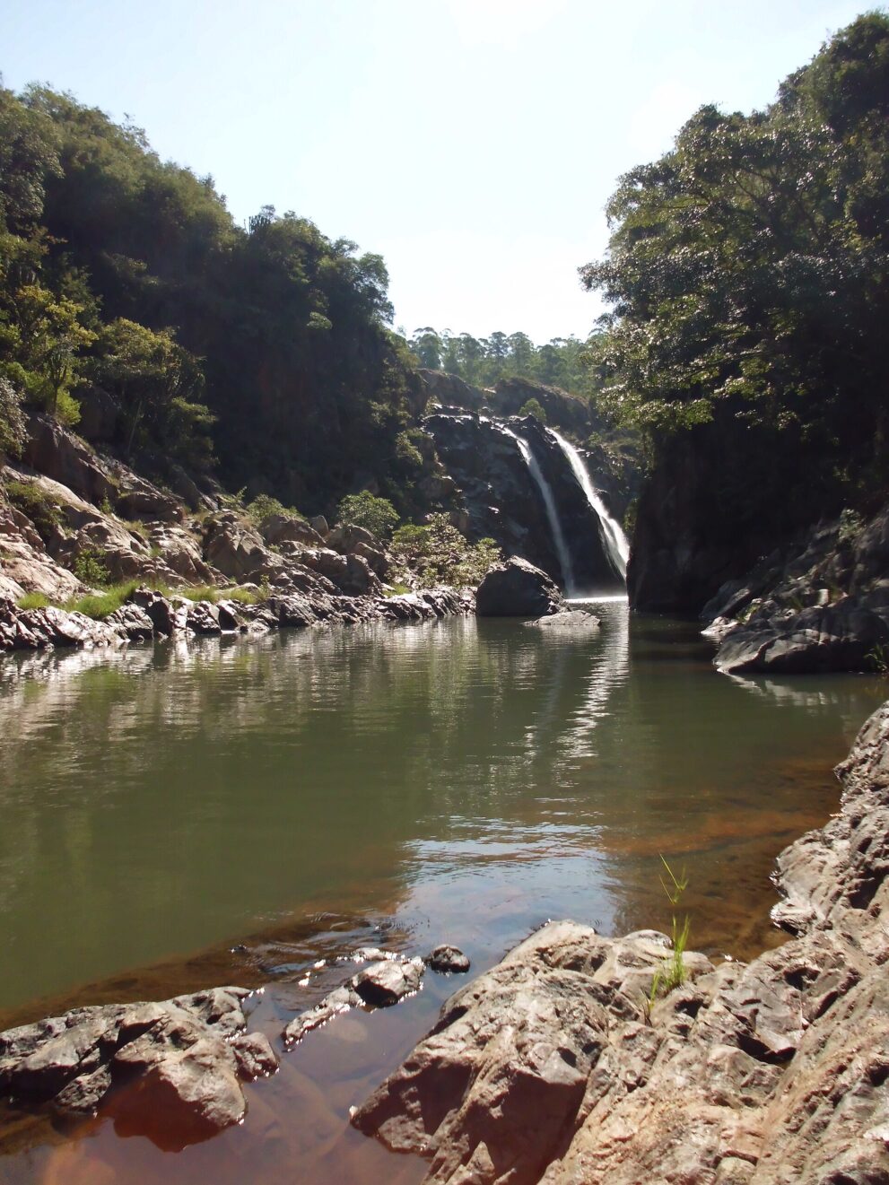 Der Wasserfall fällt zwischen Felsen und Bäumen in ein großes grünes Becken, vorn glatte Felsplatten am Ufer.