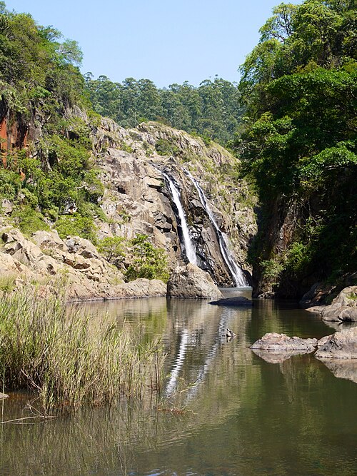 Zwei schmale Wasserstränge laufen über eine helle Felswand, unten ein spiegelndes Becken mit Schilf und Felsblöcken.