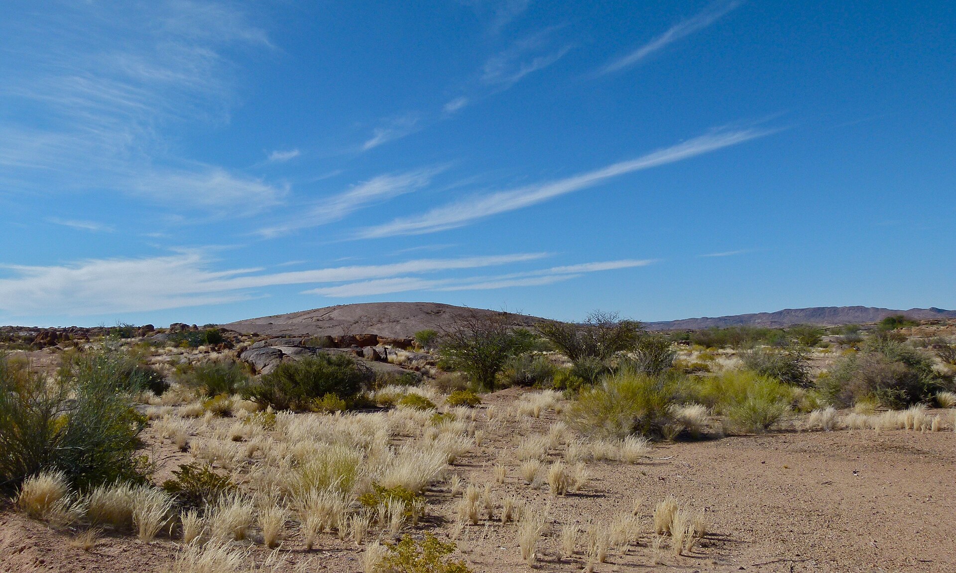 Ein flacher, kahler Felsrücken ragt über trockenes Gras und Sträucher, darüber blauer Himmel mit Federwolken, rechts eine ferne Hügelkette.