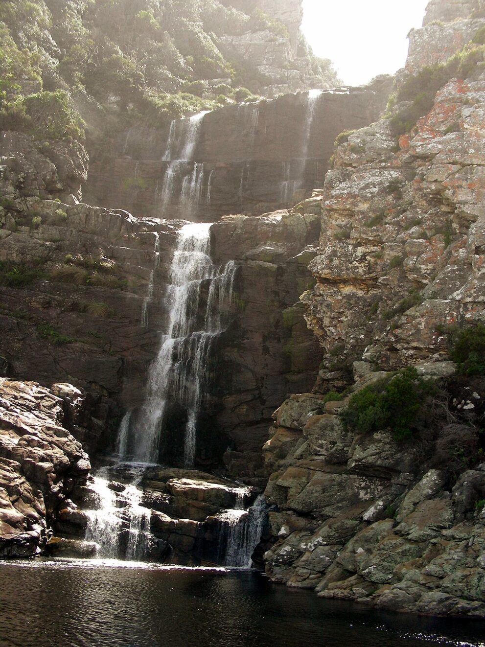 Wasserfall im Gegenlicht über Felsstufen, unten ein dunkles Becken zwischen grauen, flechtenbewachsenen Felsen
