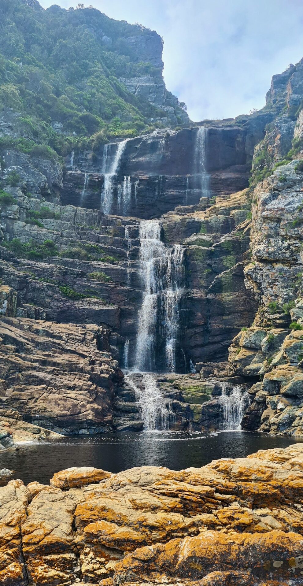 Mehrstufiger Wasserfall über dunkle Felsbänder in ein stilles Becken, vorn orange Flechten auf hellem Fels