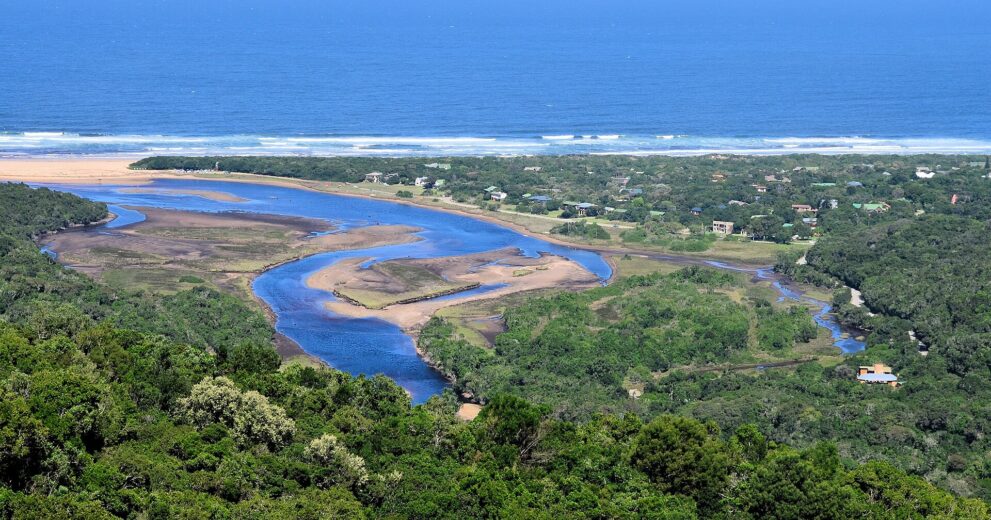 Die Lagune des Groot River windet sich mit Sandbänken durch grüne Ufer, dahinter Dorf, Strand und Meer