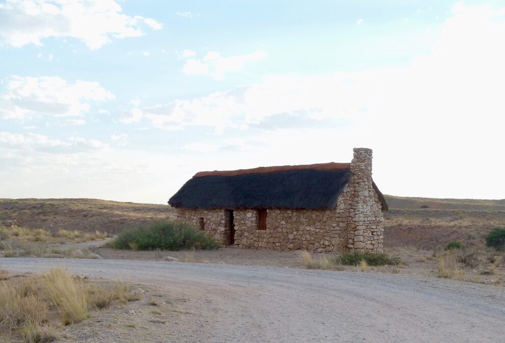 Ein kleines Haus aus hellen Kalksteinen mit Reetdach und Schornstein an einer Schotterpiste, dahinter flache Dünen