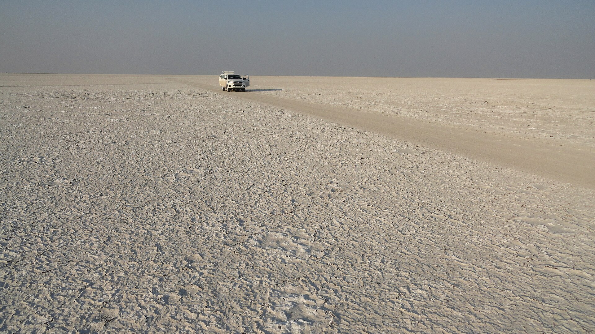 Ein Geländewagen mit offener Tür steht auf einer Fahrspur mitten in der grauweißen, rissigen Salzkruste, ringsum nur flacher Horizont.