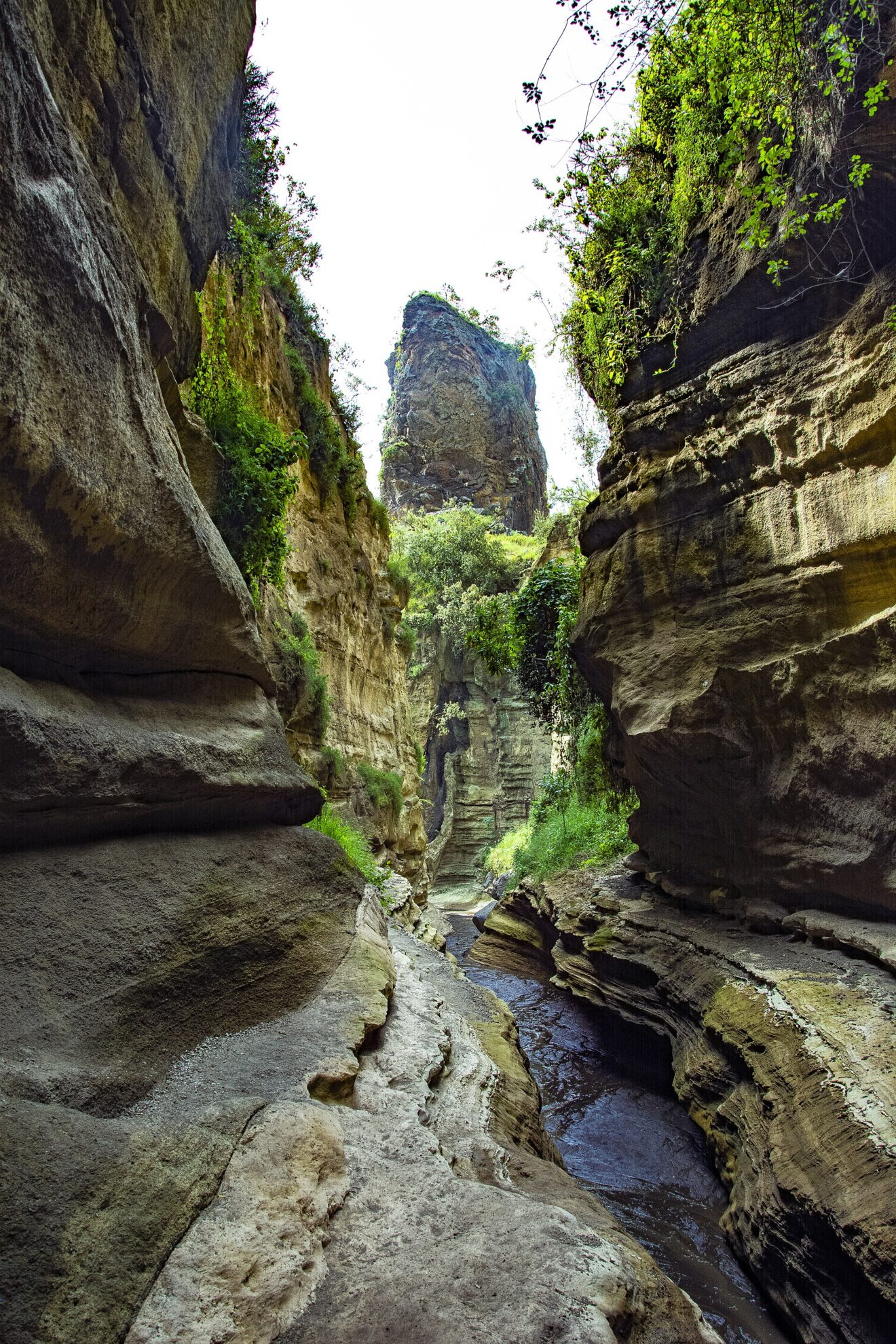 Blick durch eine enge Schlucht mit geschliffenen, gebänderten Felswänden, unten fließt flaches Wasser, im Hintergrund ragt ein Felsturm über die Kante.