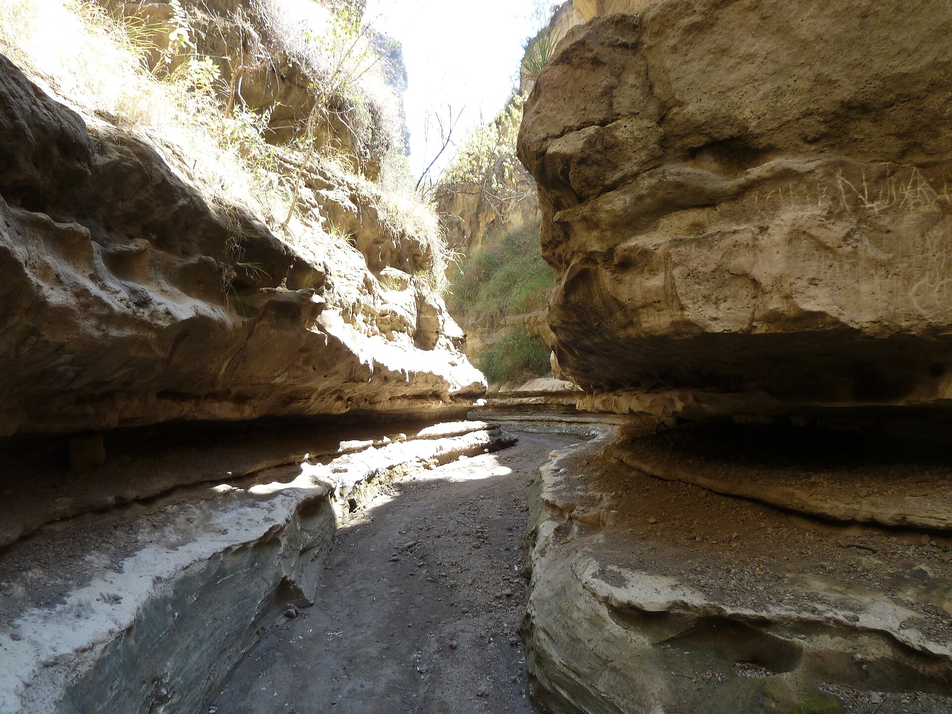 Sandiger Pfad zwischen überhängenden, ausgewaschenen Felsbänken, Sonnenlicht fällt von oben in die Schlucht.