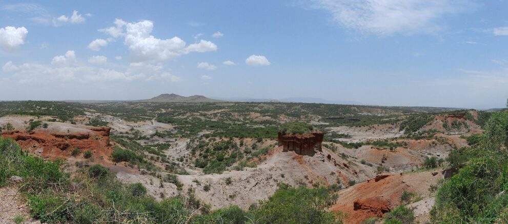 Weiter Blick in die Olduvai-Schlucht mit roten Abbrüchen links und dem Felsblock in der Mitte
