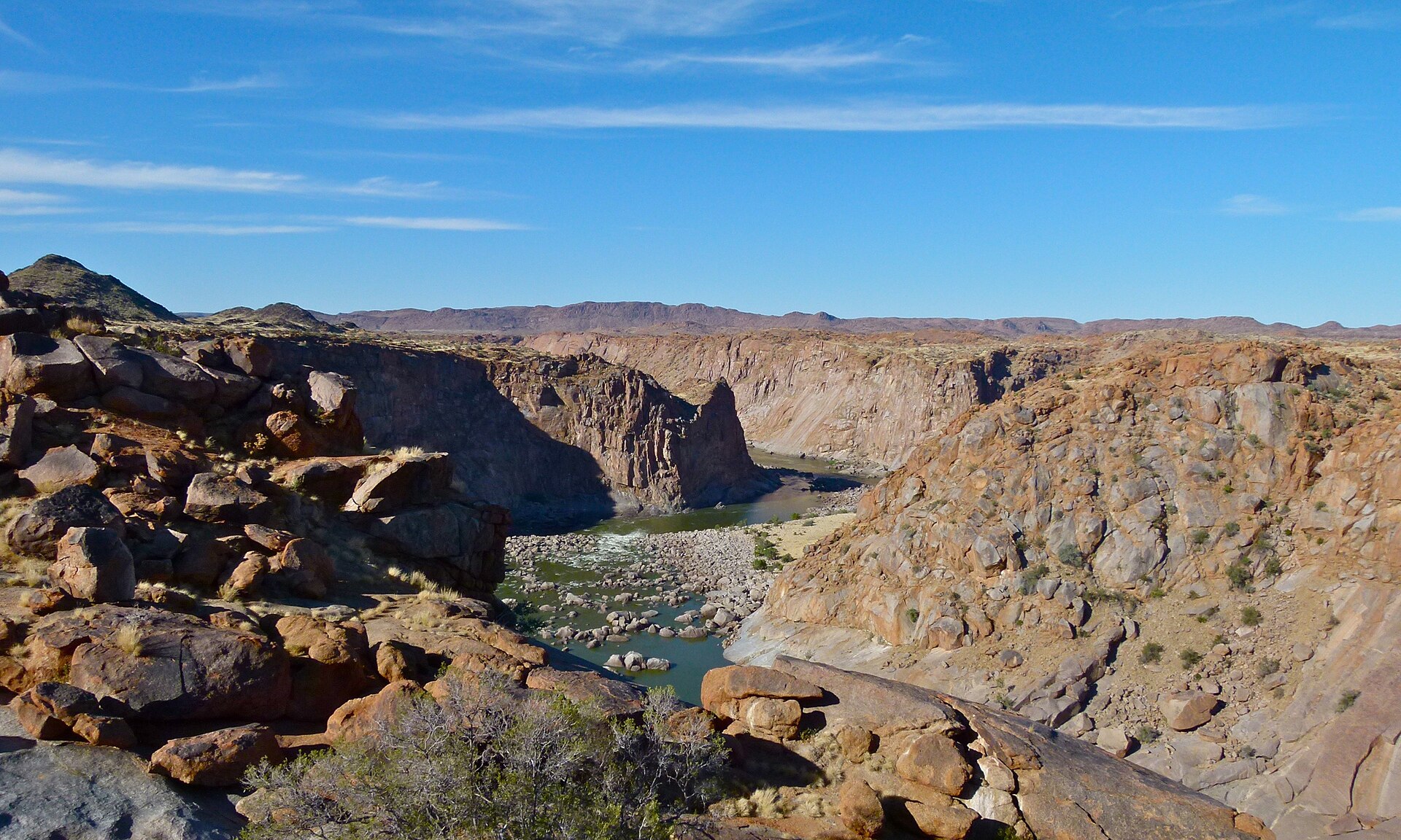 Weiter Blick über rote Granitblöcke in die Schlucht, unten Fluss und Geröll, dahinter hohe Felswände und Berge unter blauem Himmel.