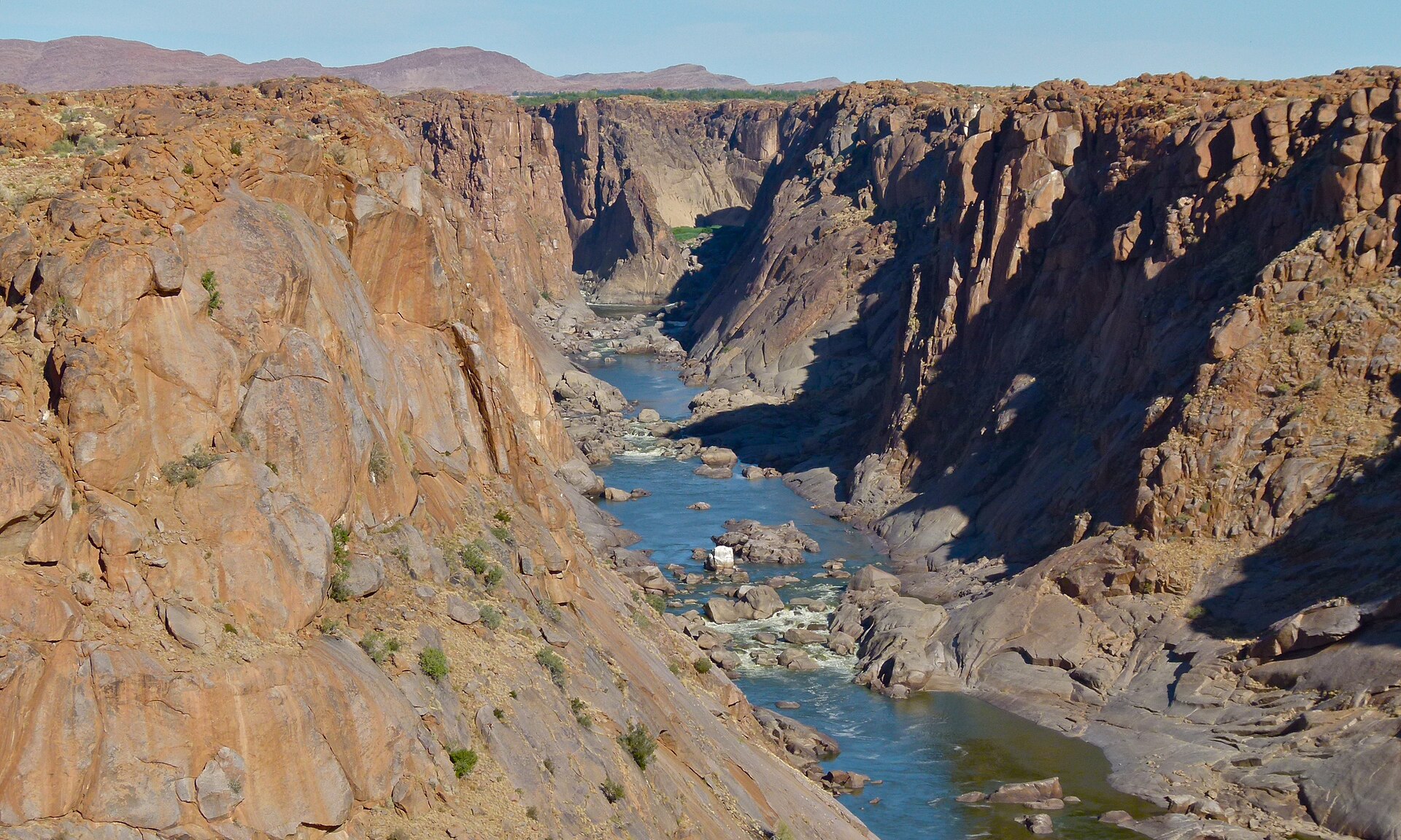 Enge, tiefe Schlucht mit senkrechten rötlichen Wänden, unten fließt der blaue Fluss zwischen Felsen und kleinen Stromschnellen.