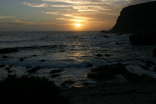 Sonnenuntergang über dem Meer, rechts eine dunkle Steilküste, vorn Felsen und Kiesstrand im Gegenlicht