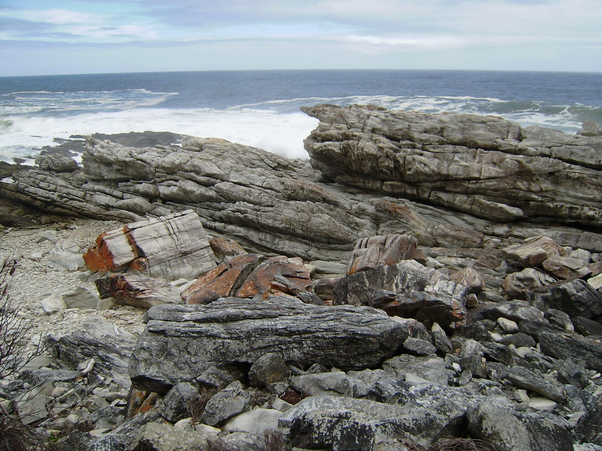 Schräg gestellte, gefaltete Felsbänke aus grauem Gestein an der Küste, dahinter Brandung und offenes Meer