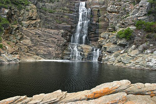 Zweistufiger Wasserfall über graue Felsstufen in ein dunkles, stilles Becken, vorn orange Flechten auf dem Fels