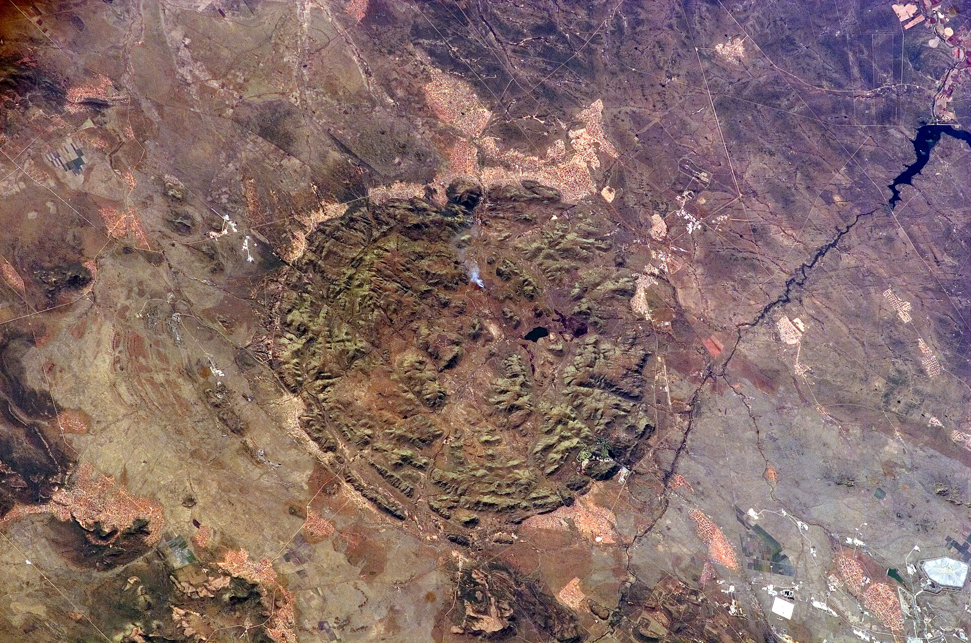 Foto aus dem All: ein runder, grünlich-brauner Hügelkomplex inmitten flacher, rötlicher Ebenen mit Straßen und Siedlungen.