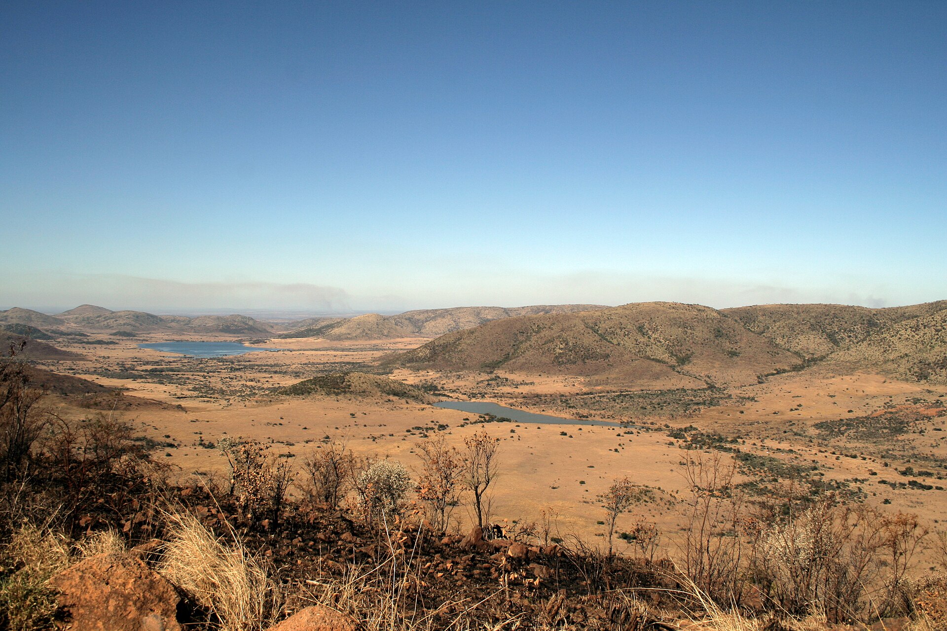 Blick von einem Felshang über trockenes, goldgelbes Grasland mit zwei Seen, dahinter flache Hügelketten im Dunst.