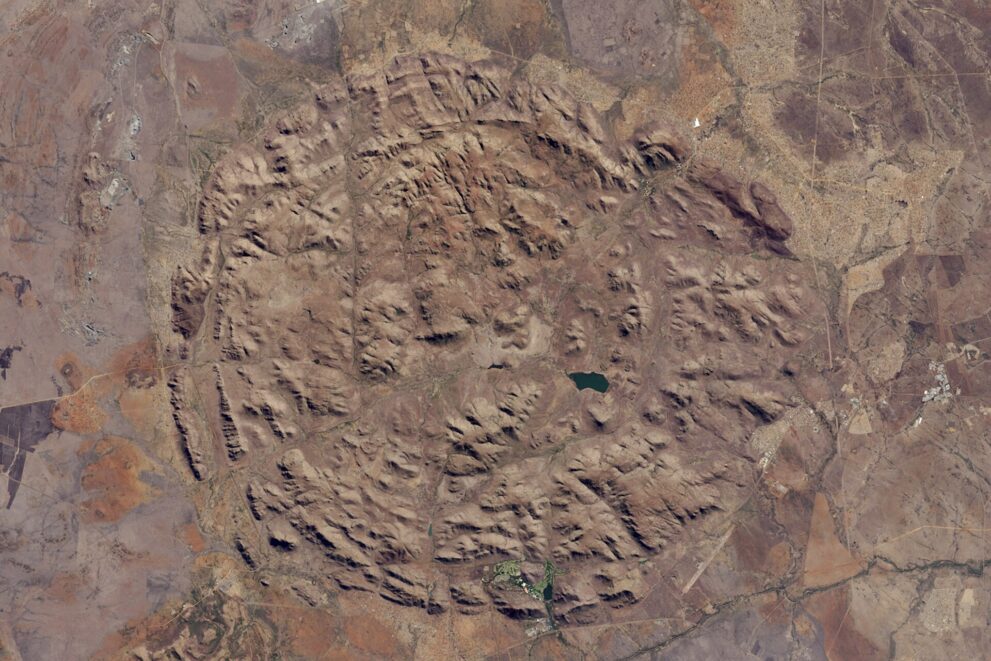 Satellitenbild: ein fast kreisrunder Komplex aus braunen Hügelketten, in der Mitte ein kleiner dunkler See, ringsum flaches Land.