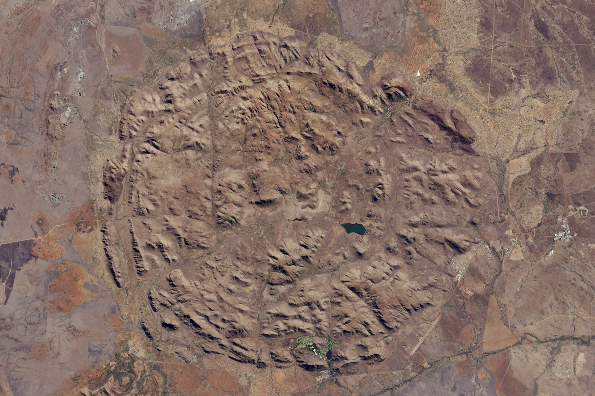 Satellitenbild: ein fast kreisrunder Komplex aus braunen Hügelketten, in der Mitte ein kleiner dunkler See, ringsum flaches Land.