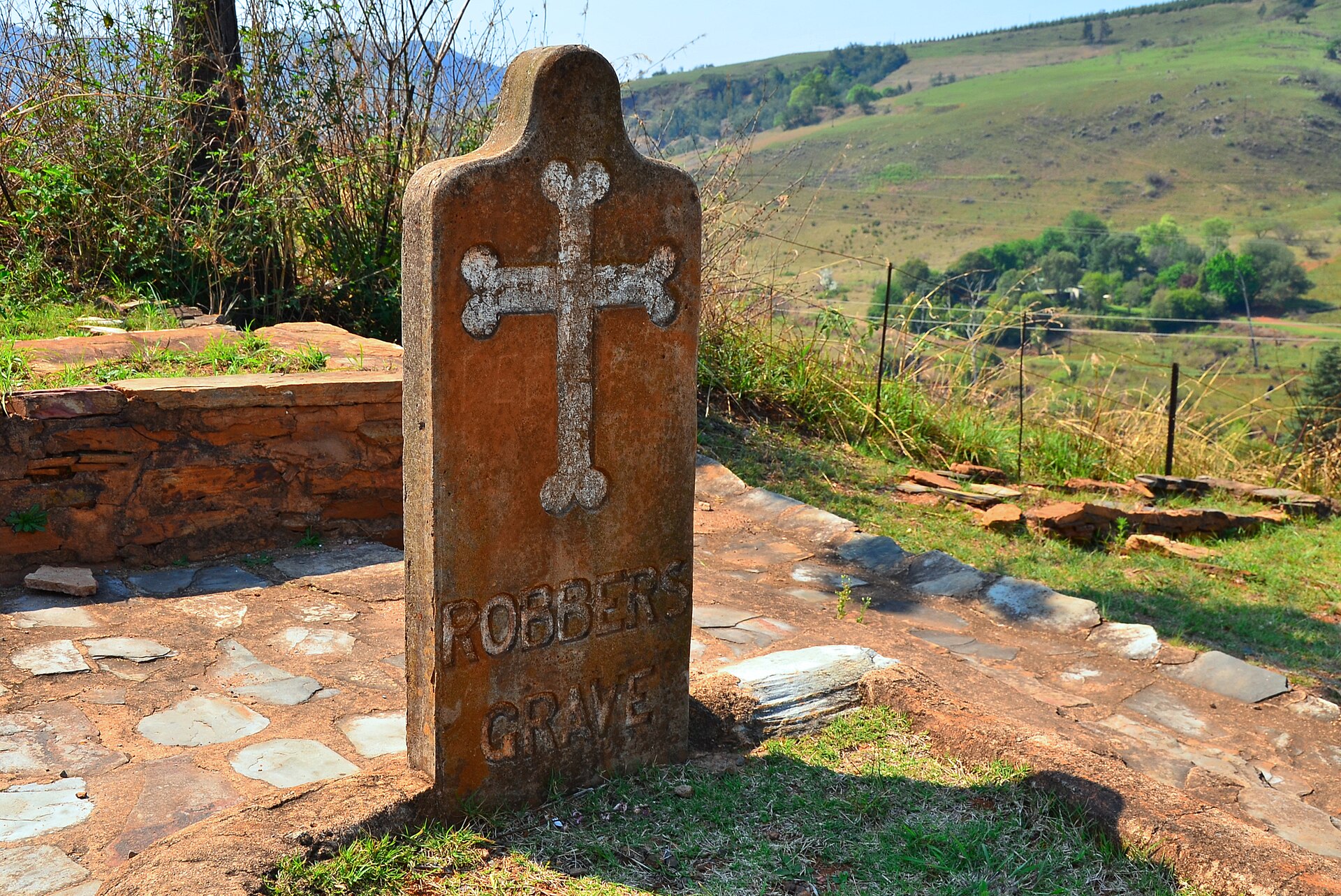 Rötlicher Grabstein mit weißem Kreuz und der Aufschrift Robbers Grave, dahinter grüne Hügel