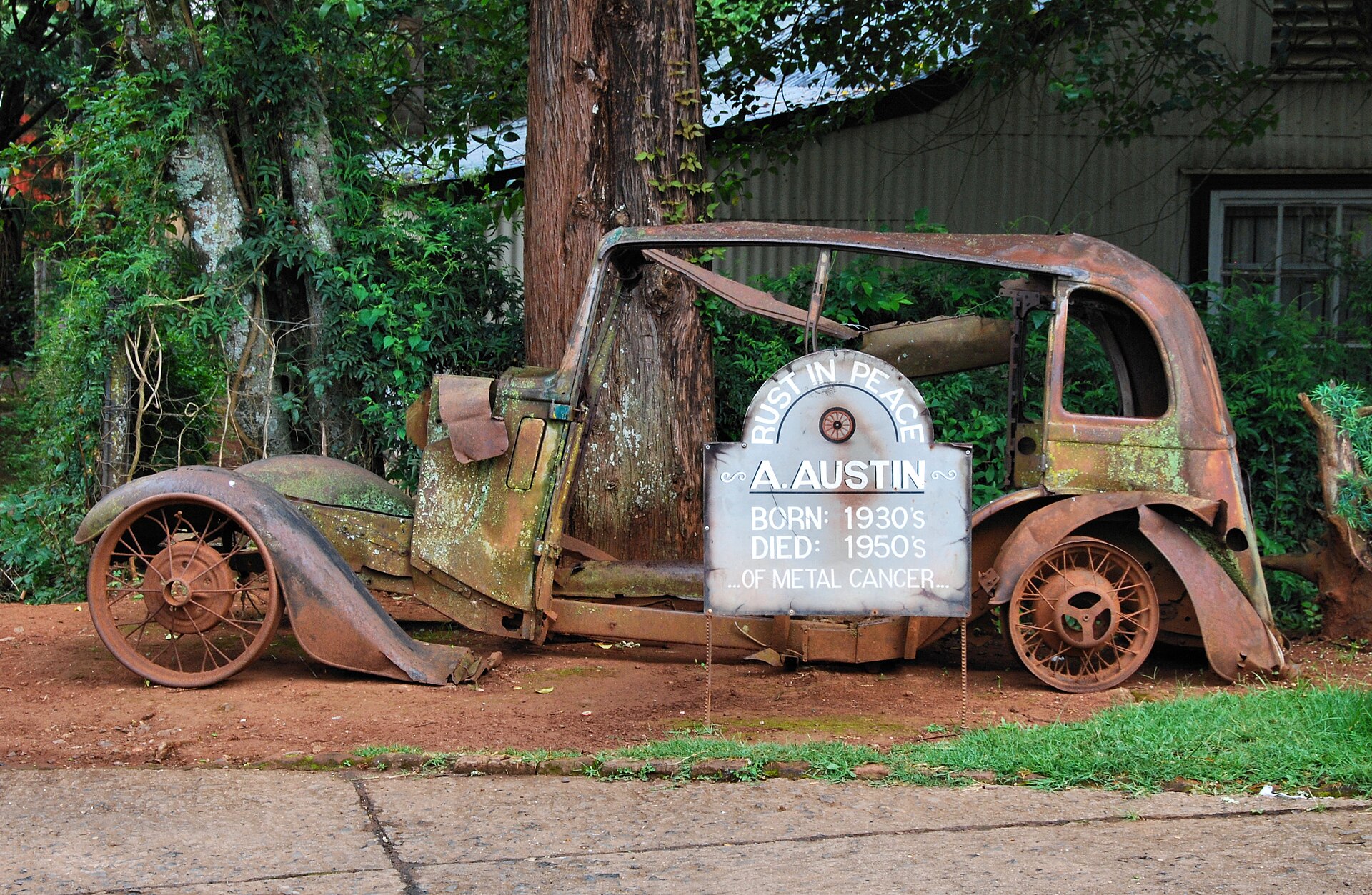 Verrostetes Autowrack vor einem Wellblechhaus, davor ein Schild mit der Aufschrift Rust in Peace