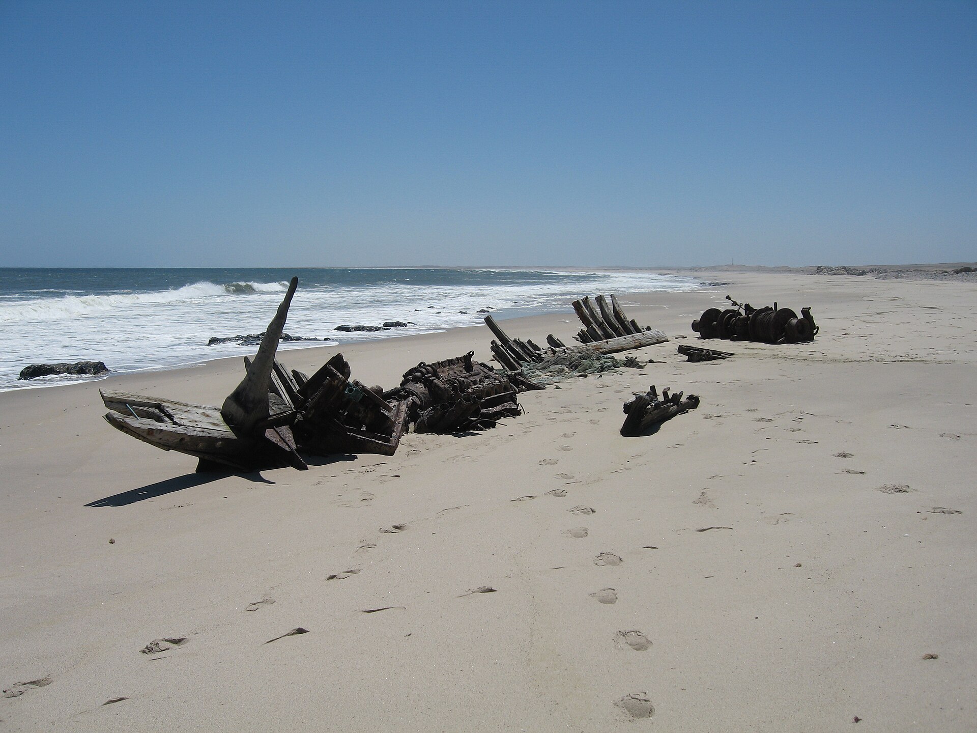 Reste eines Holzboots am Sandstrand: Bugspitze, Spanten und ein rostiger Motorblock, dahinter die Brandung