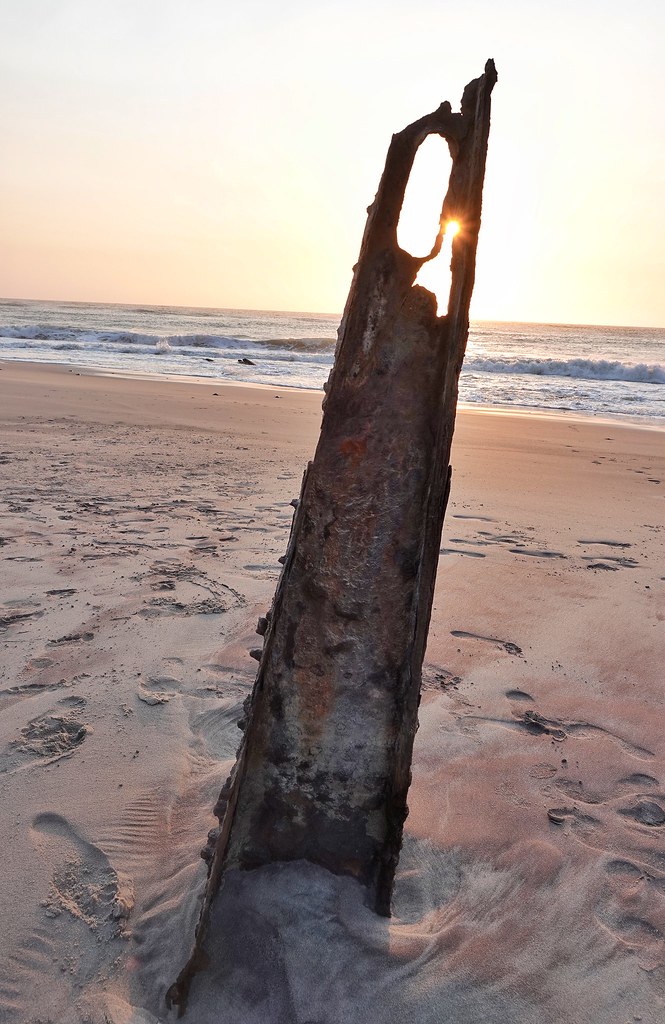 Ein aufrecht stehendes, durchgerostetes Stück Schiffsrumpf im Sand, durch ein Loch scheint die tief stehende Sonne