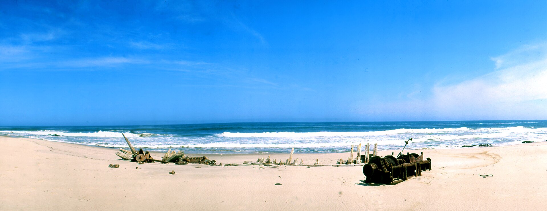 Panorama eines weiten Sandstrands mit Wrackresten, aufrechten Spanten und einer rostigen Winde vor blauem Meer